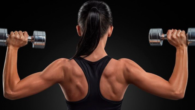 Girl standing with her back with dumbbells in each hand and showing her beautiful shoulders and arms