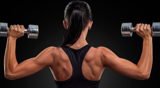 Girl standing with her back with dumbbells in each hand and showing her beautiful shoulders and arms