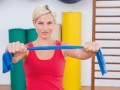 A fit girl in red undershirt is holding a blue resistance band in front of her while sitting in the gym