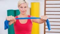 A fit girl in red undershirt is holding a blue resistance band in front of her while sitting in the gym