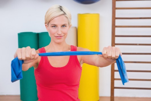 A fit girl in red undershirt is holding a blue resistance band in front of her while sitting in the gym