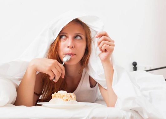 Girl with a spoon in her mouth is lying under the cover and eating a cake. Girl craving sweets because of sleep deprivation