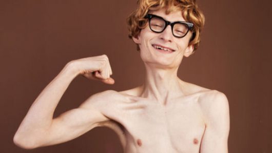 A very thin and naked guy in glasses without muscles isolated on dark flexing his biceps