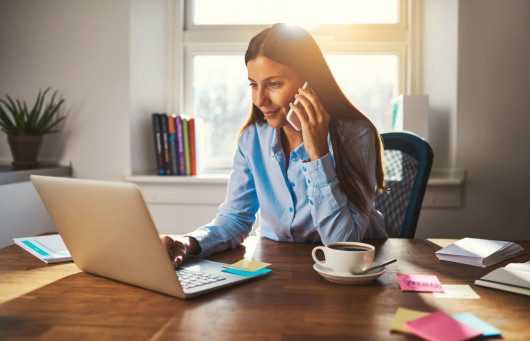 Girl is sitting at the table in front of the laptop with a cup of coffee and talking on the phone. She is working from home