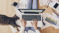 A girl with a laptop on her laps working from home. She has a cat and scattered papers around her.