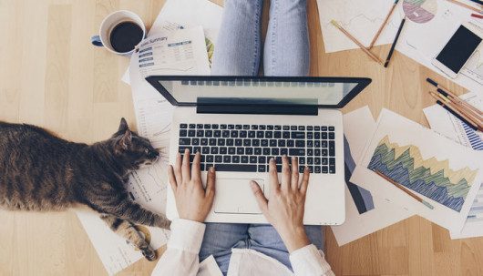 A girl with a laptop on her laps working from home. She has a cat and scattered papers around her.