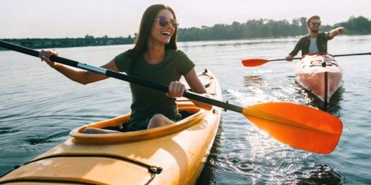 A beautiful girl and a man are kayaking on the lake in yellow kayaks. They are happy. The man in the kayak is pointing at something.