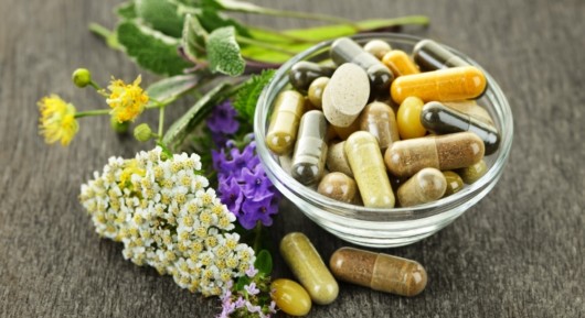 Different vitamins and dietary supplements are in the bowl on grey table with white and purple flowers