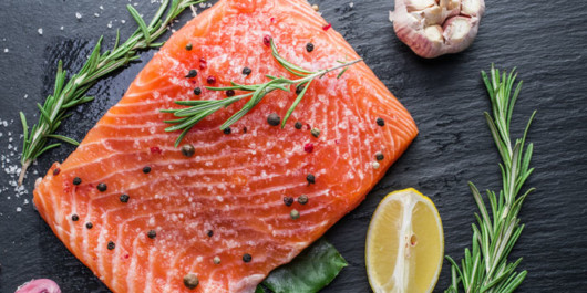 A piece of salmon with rosemary, pepper, garlic and lemon on black wooden background