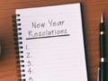A notepad where it says "new year resolutions" lies on the wooden table with a pen, sell phone and a coffee