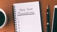 A notepad where it says "new year resolutions" lies on the wooden table with a pen, sell phone and a coffee
