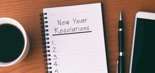 A notepad where it says "new year resolutions" lies on the wooden table with a pen, sell phone and a coffee