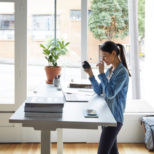 Girl in jeans shirt is is standing in front of the standing desk holding a camera and looking unto the laptop