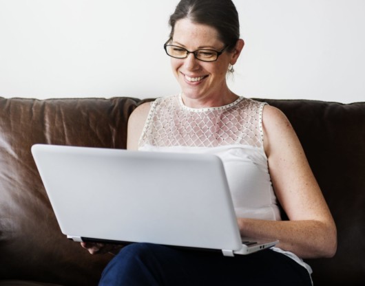 A nice girl in glasses and with a laptop is sitting on a brown sofa and smiling.