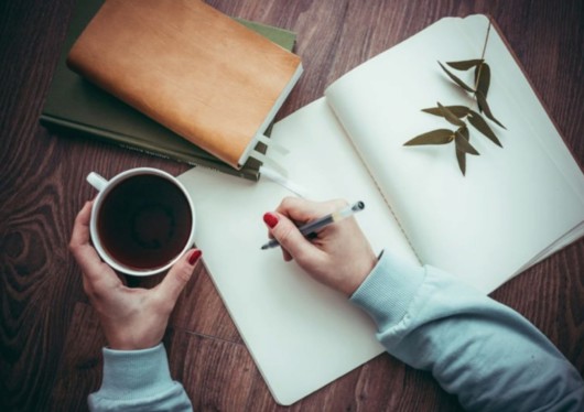 A girl is holding a pen in one hand an a cup of tea in the other one. She is going to write something in the notepad 