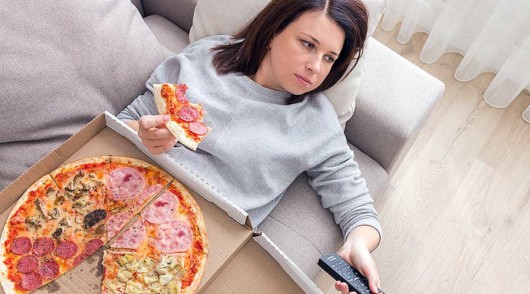 Girl with emotional eating disorder having pizza lying on the sofa and watching Tv