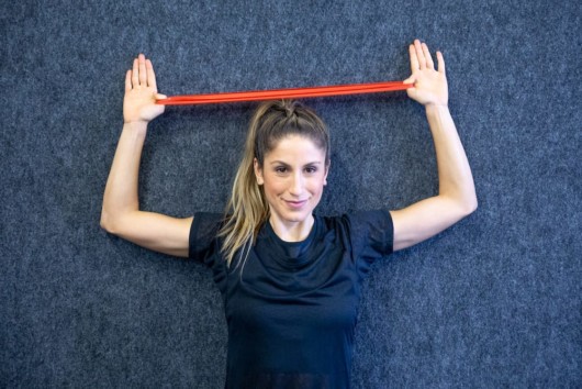 A beautiful girl in a black t-shirt on grey background is doing Resistance Band Overhead Reverse Fly .