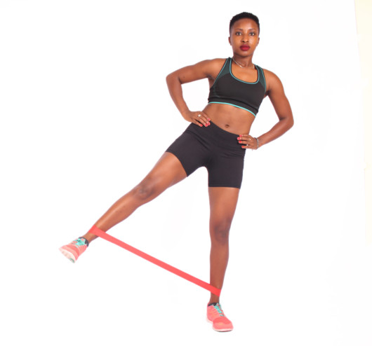 A beautiful girl in a top and shorts on white background puts her leg up to the side to shows how to do Resistance Band Side Kick