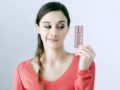 Young girl in red sweater on white background is holding birth control pills in hand and thinking if to take them or not