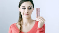 Young girl in red sweater on white background is holding birth control pills in hand and thinking if to take them or not