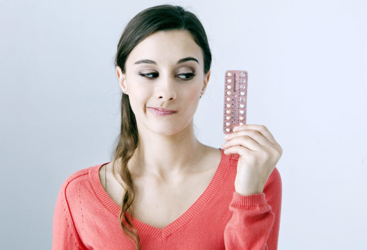 Young girl in red sweater on white background is holding birth control pills in hand and thinking if to take them or not