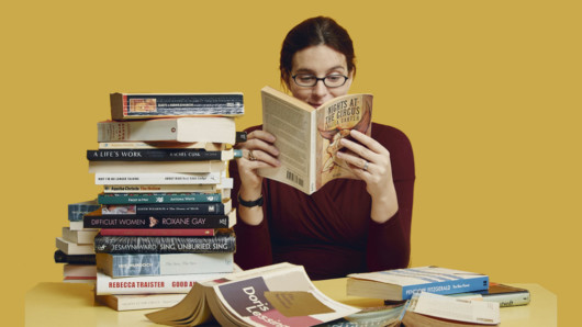 A young girl on yellow background is reading at the table with a pile of books