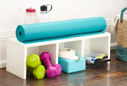 A blue fitness mat is lying on top of storage container for fitness gear. There are dumbbells, yoga brick and two water bottles