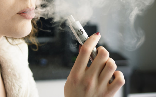 Close-up of a young woman exhaling smoke vapour from a vaping device, holding the device in her hand. Shot at home, indoors.