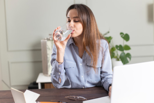 A young girl is sitting at the wooden table in front of the laptop and drinking water