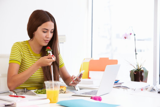 A girl is eating healthy at the table while working or studying. She eats a salad and checks her phone