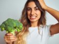 A young girl on white background is holding broccoli in her hand and is wondering what to do with it