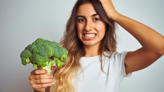 A young girl on white background is holding broccoli in her hand and is wondering what to do with it