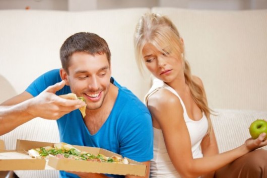 Girl with a green apple in her hand looks at the happy guy who is eating pizza