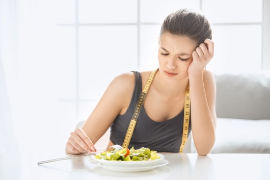 A sad girl is sitting at the white table with a measuring tape in front of the plate and not willing to eat the salad. The girl has no desire to eat the salad on the table