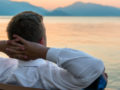 Man in white shirt is sitting with his hands behind his head on the bench overlooking the water