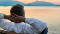 Man in white shirt is sitting with his hands behind his head on the bench overlooking the water