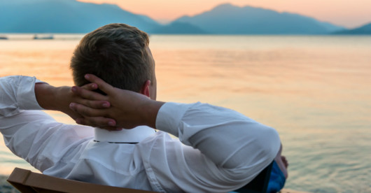 Man in white shirt is sitting with his hands behind his head on the bench overlooking the water