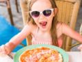 A little girl is sitting at the table with knife and fork in front of the pizza and smiling