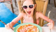 A little girl is sitting at the table with knife and fork in front of the pizza and smiling