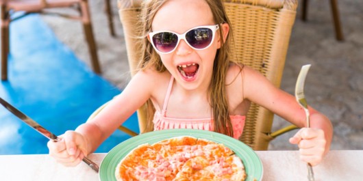 A little girl is sitting at the table with knife and fork in front of the pizza and smiling