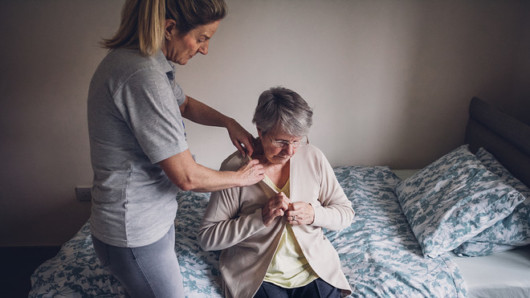 A young girl is helping to an old lady to undress. The old lady sits on the bed and trying to undress