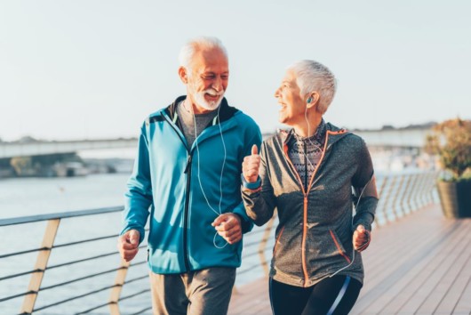 An old couple is exercising outside. An old woman and a man are running on the street and laughing. the are having fun when exercising