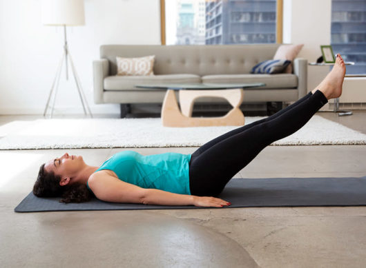 A woman in a green fitness top and black leggings is doing postpartum belly exercises at home