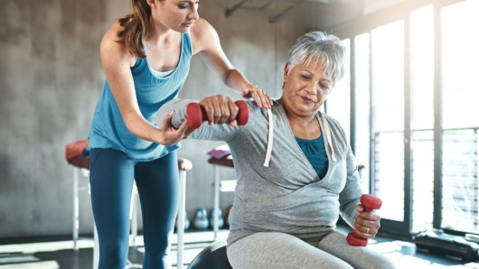 A fitness trainer is helping the old woman to do the exercise with dumbbells. The trainer is instructing the old lady in the gym