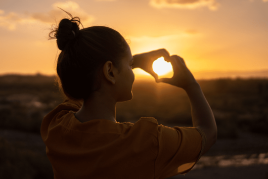 Girl is holding her fingers in the shape of heart in front of the sunset 