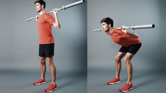 Guy in red t-shirt and black shorts on grey background with a barbell on his shoulders showing how to do good morning exercise