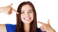 Beautiful woman in blue t-shirt pointing with her fingers at her perfect white teeth on white background