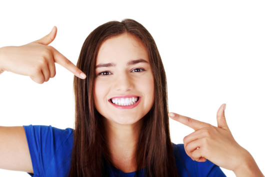 Beautiful woman in blue t-shirt pointing with her fingers at her perfect white teeth on white background