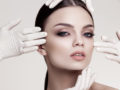 Beautiful girl's face is touched by four hands in white medicine gloves on grey background