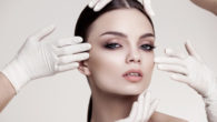 Beautiful girl's face is touched by four hands in white medicine gloves on grey background